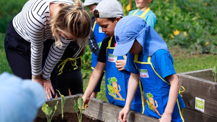 Zwei Kita-Kinder stehen mit einer Kindergärtnerin am Hochbeet bei 