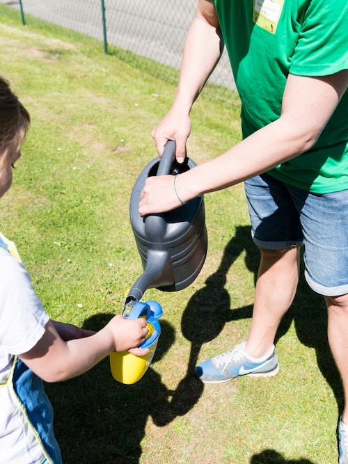 Eine kleine blau-gelbe Kinder-Gießkanne wird aus einer großen Gießkanne aufgefüllt bei 