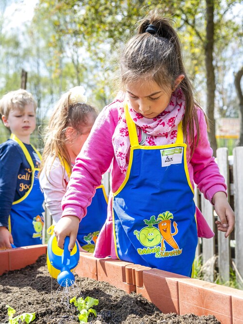 Kita-Kinder stehen um ein Hochbeet herum, ein Mädchen gießt die frisch gepflanzten Setzlinge bei 