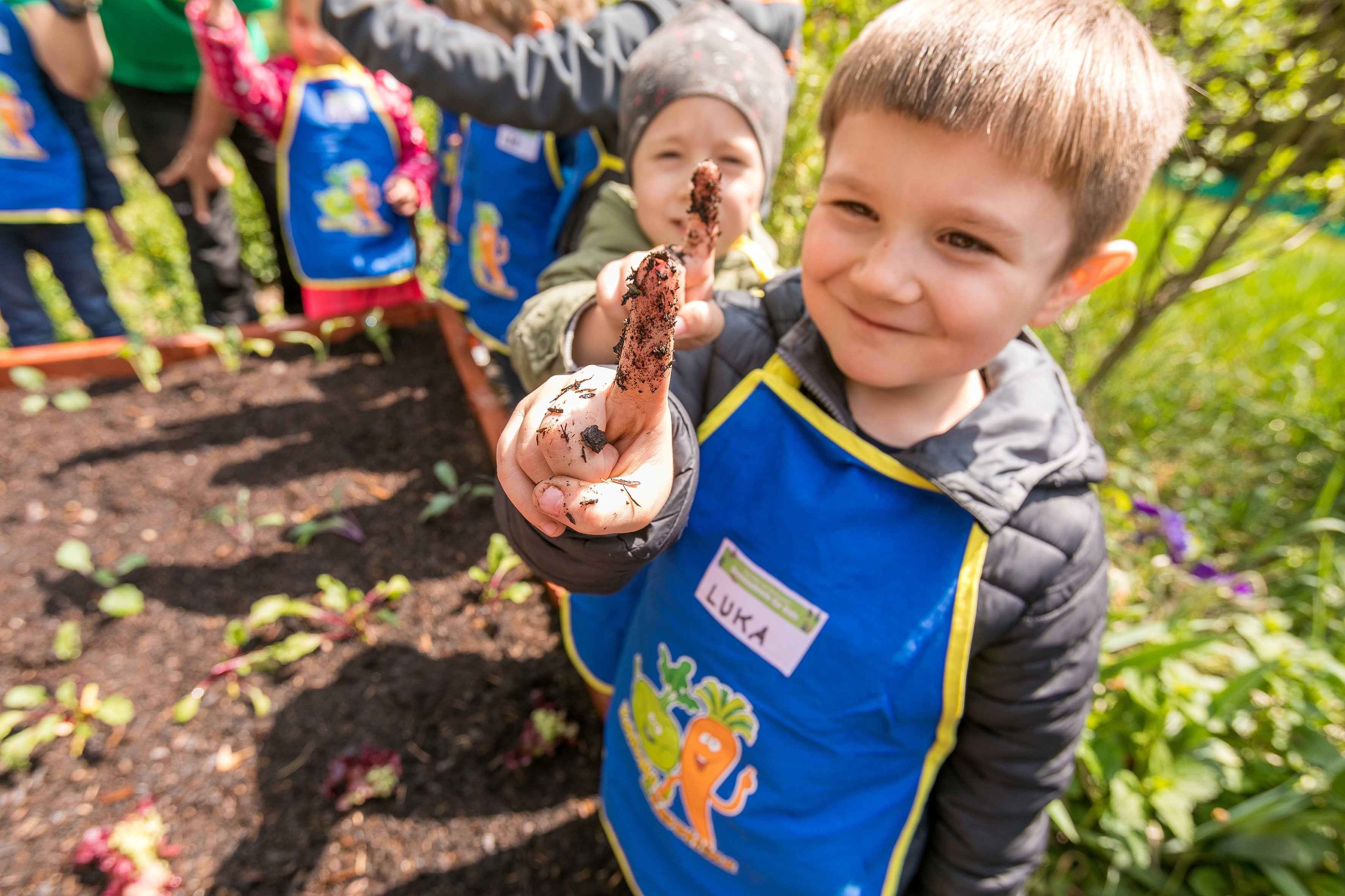 Kita-Kinder am Hochbeet bei "Gemüsebeete für Kids". Ein Junge zeigt einen Finger hoch,mit dem er zuvor die Erde getestet hat.