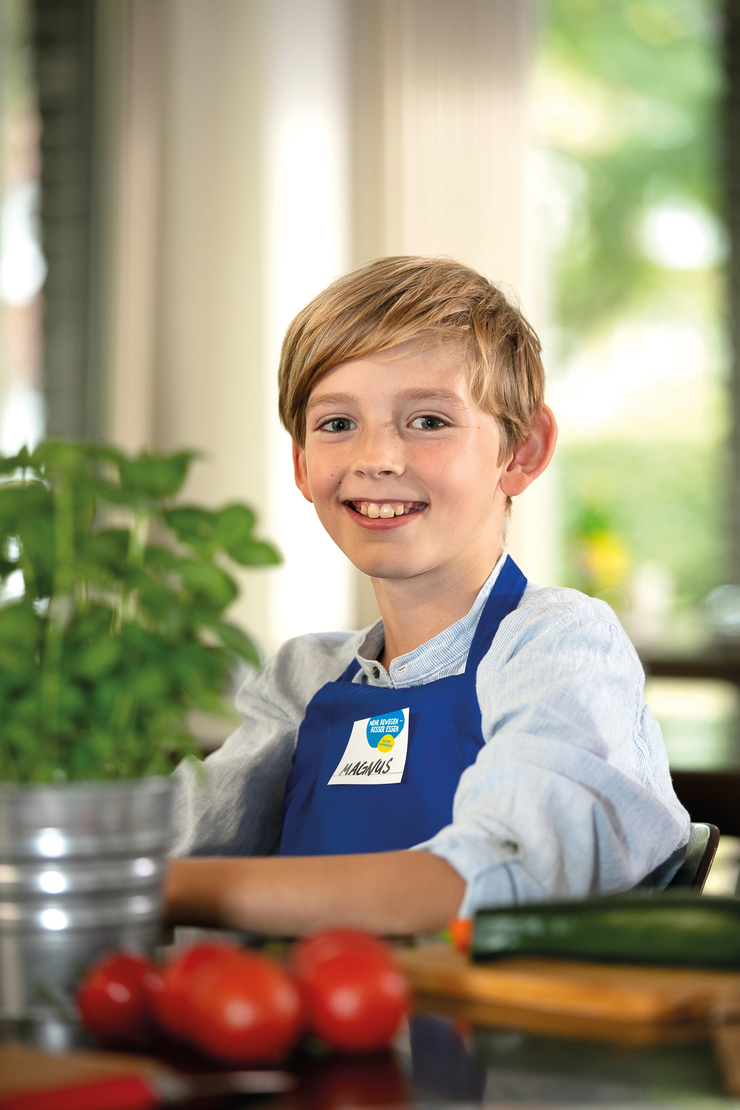Ein Junge in einer blauen Schürze lächelt in die Kamera beim Projekttag "Mehr bewegen - besser essen" der EDEKA Stiftung.